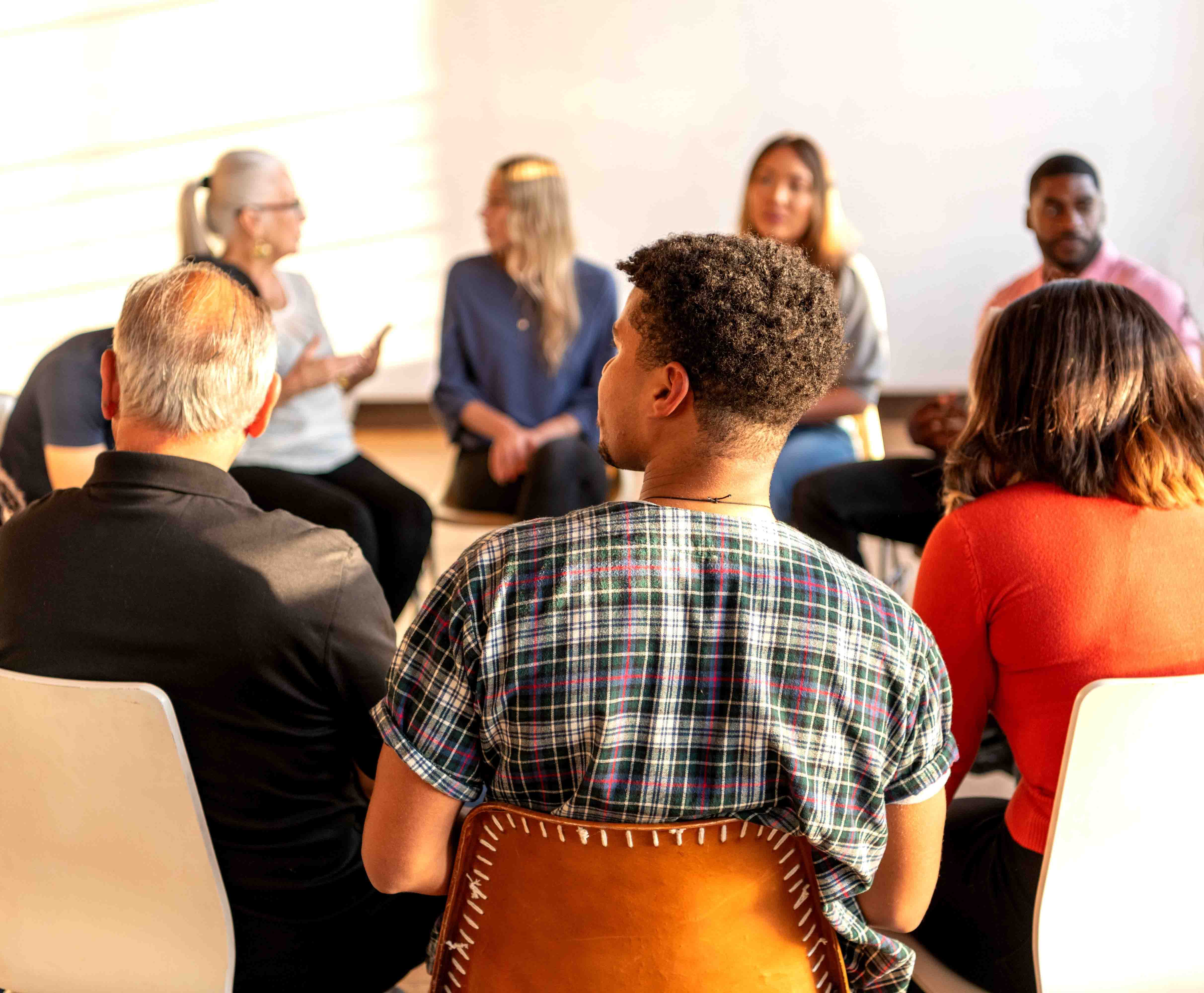 A diverse group of people sit in a circle, engaged in conversation. The setting is bright and casual, suggesting a workshop or group discussion.